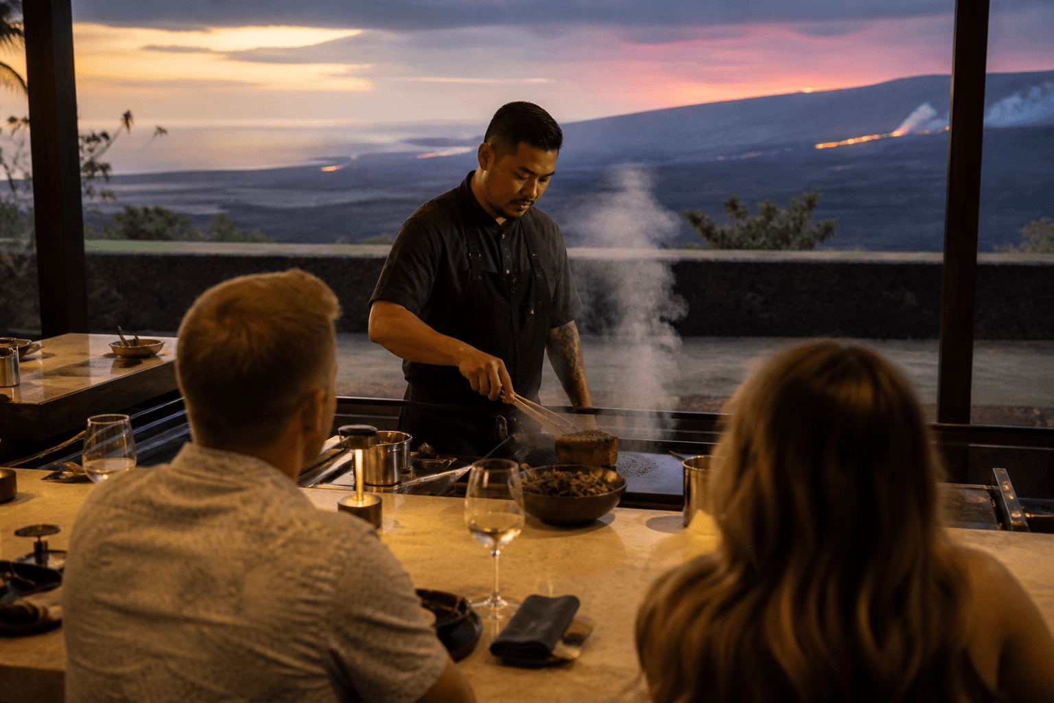 Guests at a Kona kitchen counter at dusk watching a sear, lava beyond.