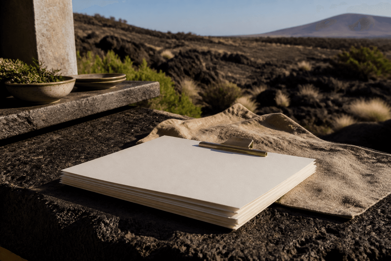 Unused ivory papers under a brass clip on a Kohala lava-stone pass in hard sun.