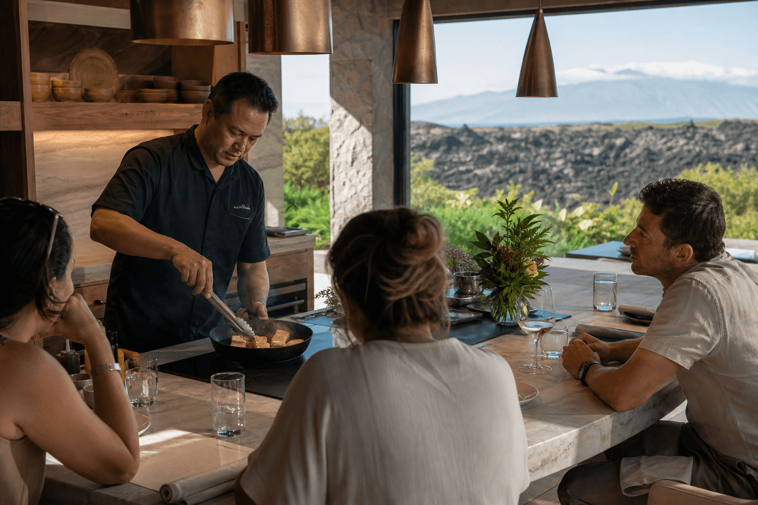 Guests at a Kona kitchen counter watching a sear, lava and coffee slopes beyond.